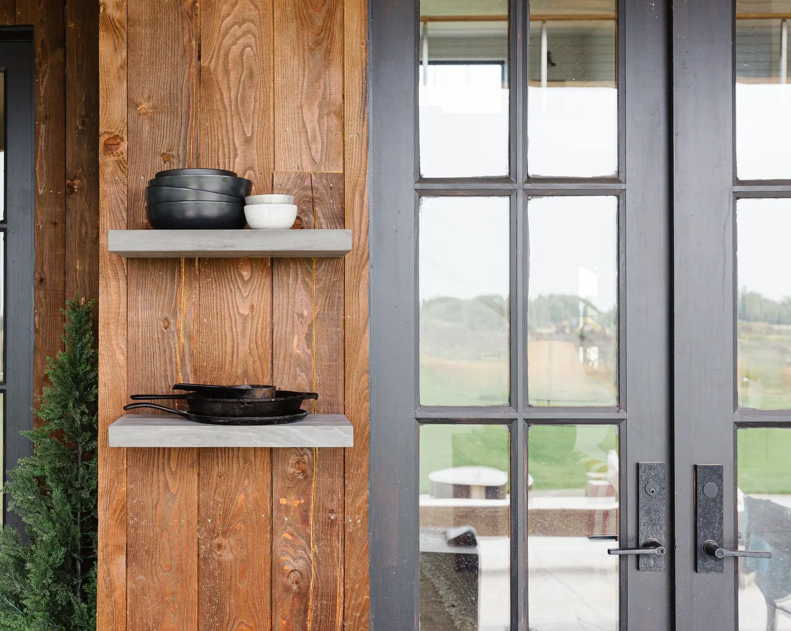 A textured wood-clad wall with charcoal floating shelves and black ceramics, glass doors opening to the prairie. Photo from ultrashelf.com.