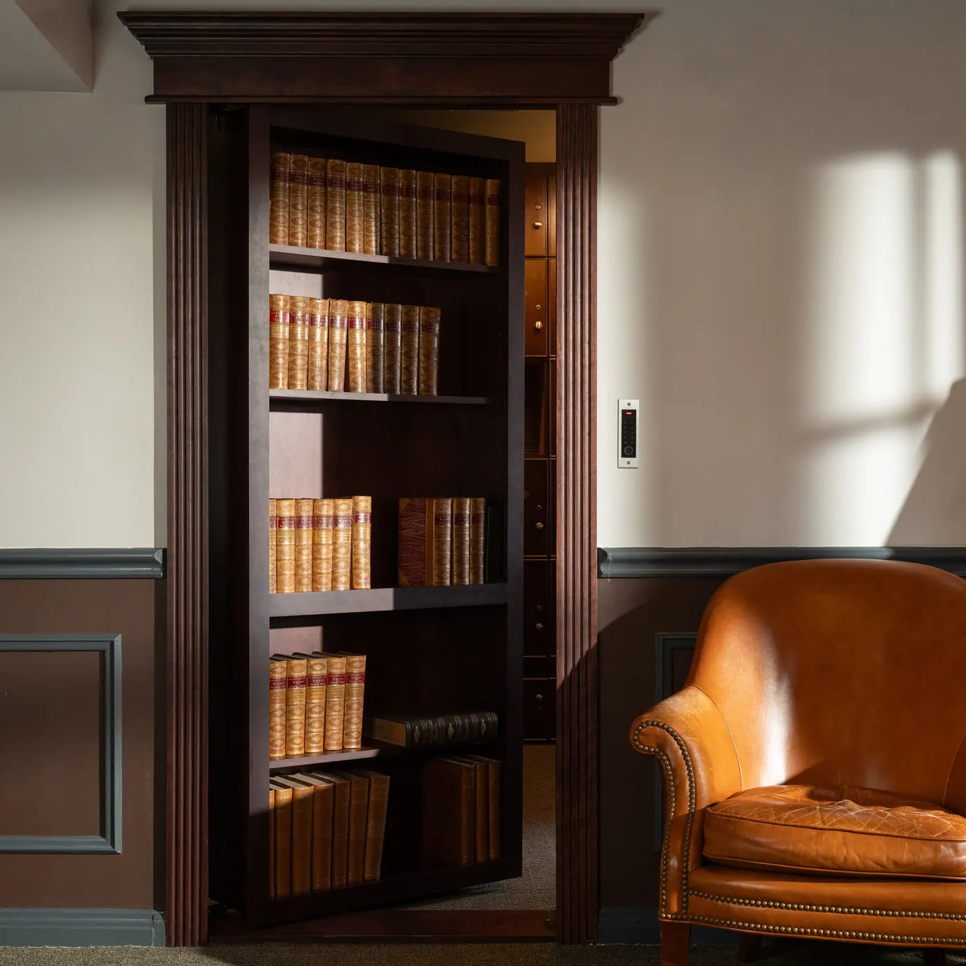 A dark walnut Murphy Door® hidden bookcase door standing open in a warm library with a leather chair. Photo from murphydoor.com.